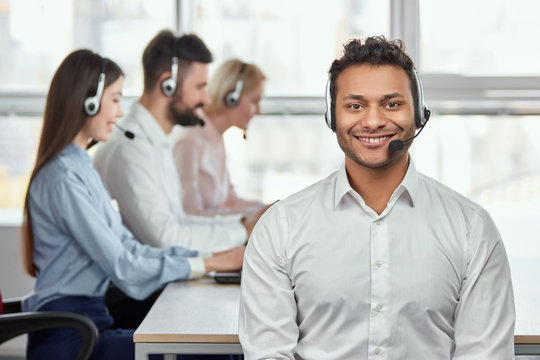 Young friendly male customer service operator. Portrait of a smiling businessman with headsets in a call centre. Bright windows background.