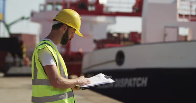 A dock worker standing at the harbor amidst shipping industry activity while taking a call on his mobile. Shot on RED Epic.