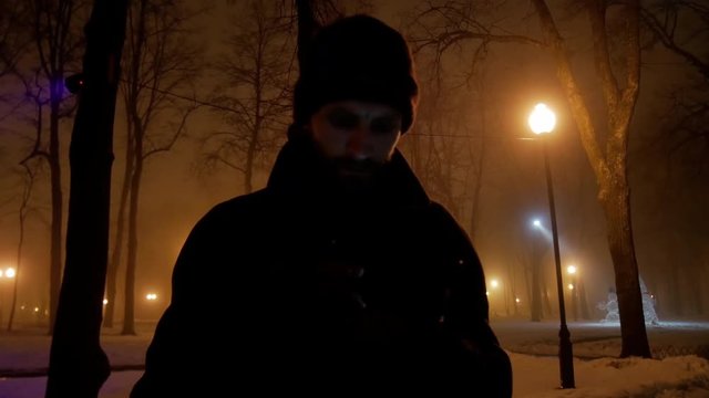 4k Bearded brutal man with mobile phone typing messages in dark winter fog danger park lowlight 