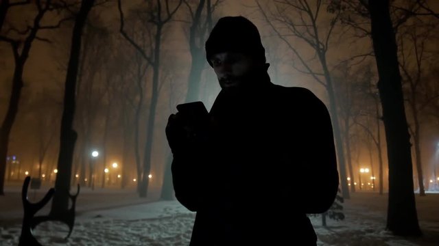 4k Bearded danger criminal look man with mobile phone in foggy night city park reads messages