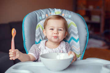 Portrait of cute adorable Caucasian child kid girl sitting in high chair eating cereal with spoon. Everyday lifestyle. Candid real authentic moment