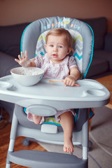 Portrait of cute adorable Caucasian child kid girl sitting in high chair eating cereal with spoon. Everyday lifestyle. Candid real authentic moment