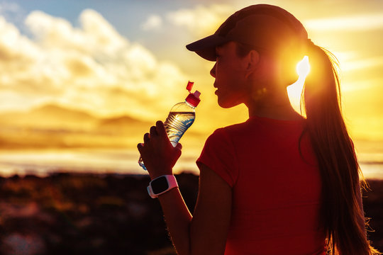 Fitness Woman Drinking Water From Sports Bottle On Afternoon Workout After Run Training Jogging Outdoors At Sunset. Girl Wearing Running Cap Silhouette Against Sun Flare.