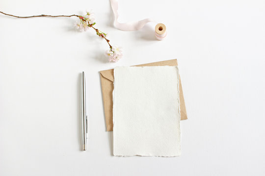 Wedding spring styled stock photo. Feminine desktop mockup with blossoming cherry tree branch, silver ballpoint pen, craft envelope and blank greeting card, white background. Empty space. Top view.