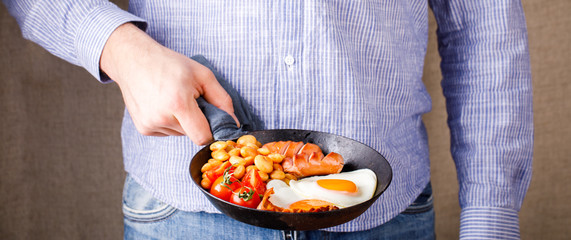 Traditional English Breakfast in the Frying Pan .Egg in the Heart shape. Valentine's Day. Festive Food in the hands of a young man Background.Sausages, Bacon, Beans,Orange juice