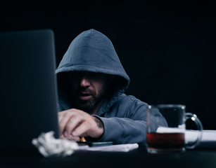 male hacker in a sweatshirt with a hood sits behind a laptop