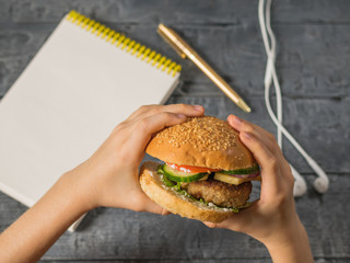 The girl holds a freshly prepared Burger over a table with a pen, a notebook and headphones.