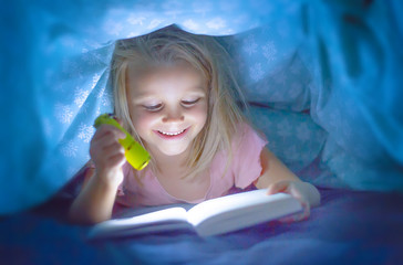 sweet beautiful and pretty little blond child girl 6 to 8 years old lying under bed covers reading book in the dark with torch flashlight smiling happy