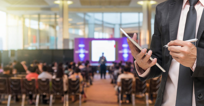 Businessman using the tablet on the Microphone over the Abstract blurred photo of conference hall or seminar room with Speakers on the stage and attendee background, Business meeting concept