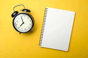 black alarm clock with blank white notebook on yellow background color