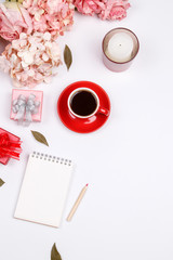 Valentines day background. Feminine desk workspace frame with pink beige floral bouquet, red coffee cup, candle, gift box and notebook on white background with copy space. Flat lay. Top view.