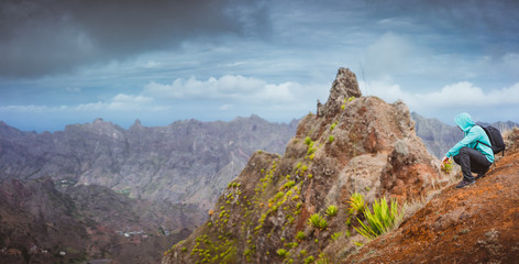 Man hiker with backpack sitting on the mountain top and looking down to the valley. Stunning arid landscape on Santo Antao island, Cabo Verde