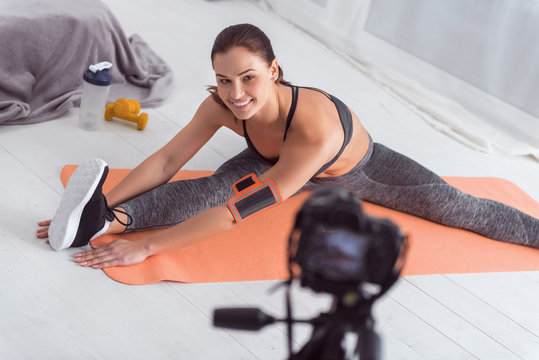 Exercising hard. Beautiful inspired fit dark-haired young woman smiling and stretching while sitting on the carpet and making a video for her blog