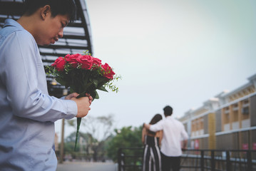 heartbroken man holding bouquet of red roses feeling sad while seeing woman dating with another man. broken heart in love concept