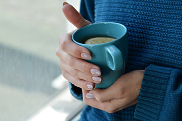 Woman in knitted sweater holding a cup of hot tea with lemon in hands with manicured nails. Turquoise and blue colors