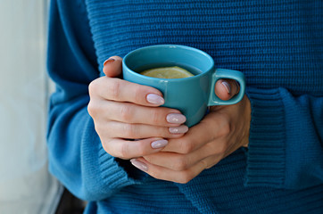 Woman in knitted sweater holding a cup of hot tea with lemon in hands with manicured nails. Turquoise and blue colors