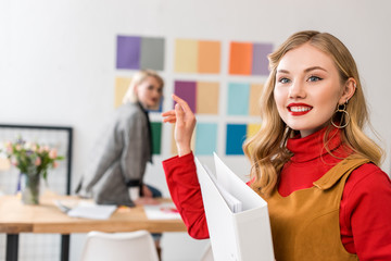 smiling magazine editor with folder in modern office with colleague and color palette on wall behind