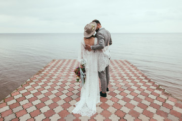 rear view of bride and groom in boho style hugging on pier at lake