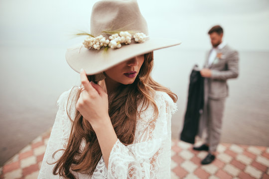 Beautiful Bride In Wedding Dress And Hat In Boho Style On Pier At Lake, Groom Standing Behind