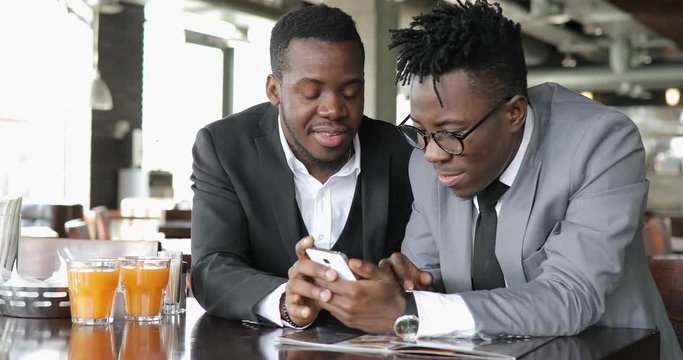 African businessmen friends discussing funny photos together on smart phone in cafe