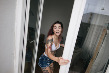 Fototapeta na wymiar surprised young woman in bra and denim shorts looking at camera through open window