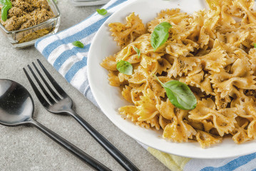 Vegan dinner. Farfalle pasta with pesto sauce and basil leaves on white plate, grey stone background, copy space