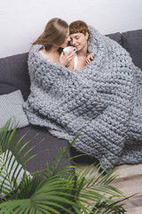 happy lesbian couple with cup of coffee embracing under knitted wool blanket on couch
