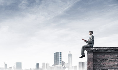 Man on roof edge reading book and cityscape at background