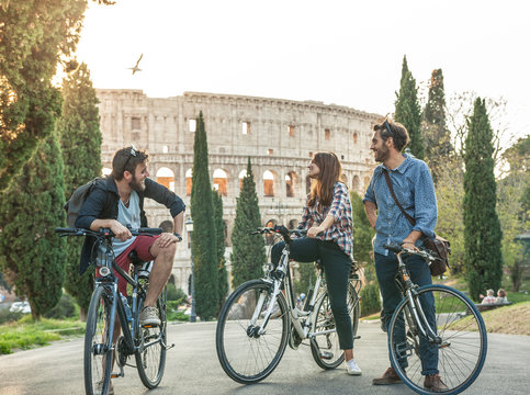 Three young friends tourists with bikes in colle oppio park in front of colosseum on road with trees at sunset having fun talking laughing in Rome lens flare