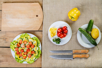 On a wooden background there are knives, a plate with salad, cucumber, tomato and pepper
