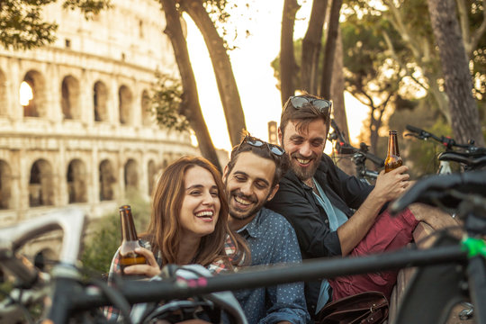 Three young friends tourists with bikes sitting on bench in front of colosseum under tree at sunset drinking beers having fun in Rome