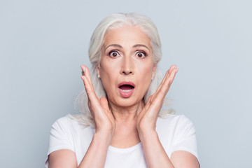 Pretty, nice, old, shocked, scared woman with wide open eyes and mouth in t-shirt holding palms near cheeks looking at camera over gray background