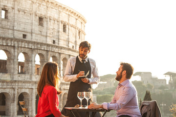 Elegant waiter writing orders on notebook serving a young happy couple in bar restaurant in front of colosseum in rome at sunset