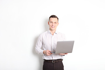 Smiling confident young man, no tie, holding grey laptop device and typing while standing against solid white wall. Wireless internet, wifi, enter password. Shaved, no facial hair. Isolated background