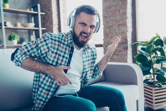 Attractive, bearded man sitting on the couch in living room, having headphones on his head, listening his favorite music, singing a song dreaming like playing guitar