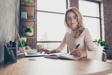 Portrait of beautiful confident with blonde curly hair clever woman wearing casual clothes, she is sitting at the table in front of computer and checking information in her diary and on the internet