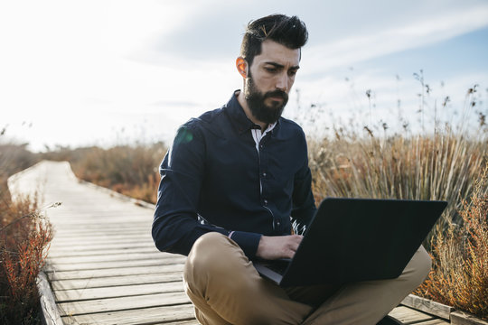 Casual man with laptop in natureConcentrated man relaxing on wooden pathway in field browsing laptop in sunlight.