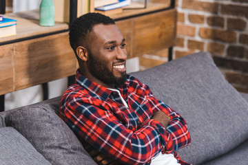 Obraz premium smiling young african american man sitting with crossed arms on sofa and looking away