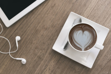 Top view A cup of hot coffee with heart shaped latte art with tablet and earphone on wooden background.