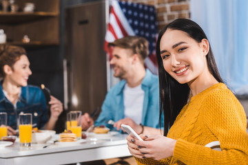 young asian woman using smartphone and smiling at camera while friends eating behind