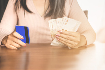 Asian woman sitting at a table choosing between credit card and bank notes for payment or preparing money for debt