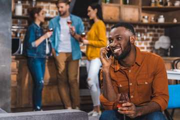 smiling african american man talking by smartphone while friends drinking wine behind