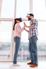 First try. Low angle of young positive nice couple taking each other hands while using VR glasses and standing near window