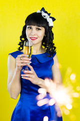 A girl with a glass of champagne smiles in front of the camera.. Beautiful brunette girl in a blue dress drinking champagne on a yellow background