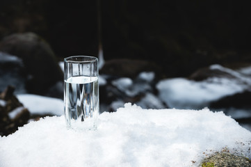 Naklejka na ściany i meble A transparent glass glass with drinking mountain water stands in the snow against a background of a clean frost mountain river in winter.