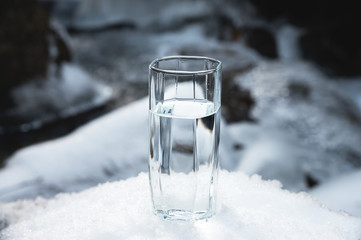 A transparent glass glass with drinking mountain water stands in the snow against a background of a clean frost mountain river in winter.