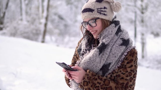 Attractive girl wears glasses typing message by smartphone in winter snowy park