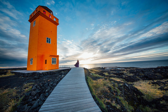 Abstract photo of woman act silhouette in dress in golden sunset near big orange lighthouse in Iceland.Fashion and nature concept artistic panorama.Western Icelandic untamed nature.Freedom