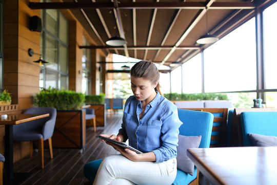 Young girl using tablet at restaurant   and sitting in on blue sofa. Pretty woman wears jeans shirt and has pony tail. Concept of modern gadgets and hotspot Internet.
