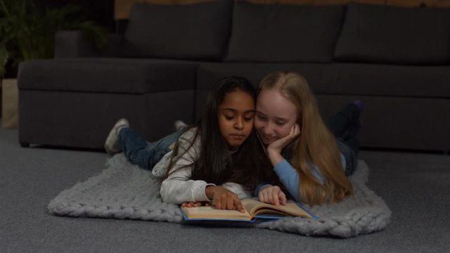 Smiling sweet multi ethnic kids reading fairytales and lauging while lying on the floor in living room. Joyful little girls spending leisure together at home and reading a book.